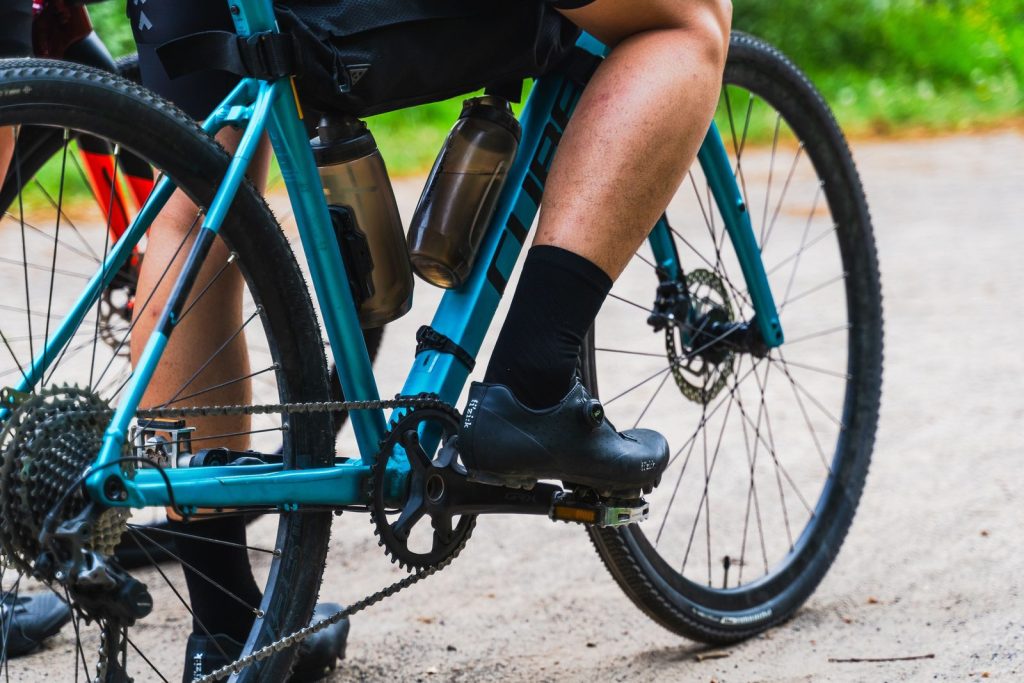 Close-up of a cyclist pedaling a teal gravel bike with bottles on the frame along a dirt path.