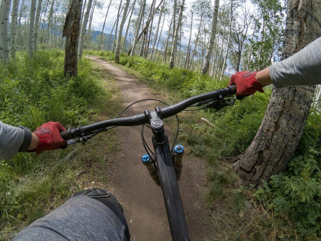 Rider's-eye view from a mountain bike on narrow dirt singletrack through an aspen grove near Aspen, Colorado, with mountains in the distance.
