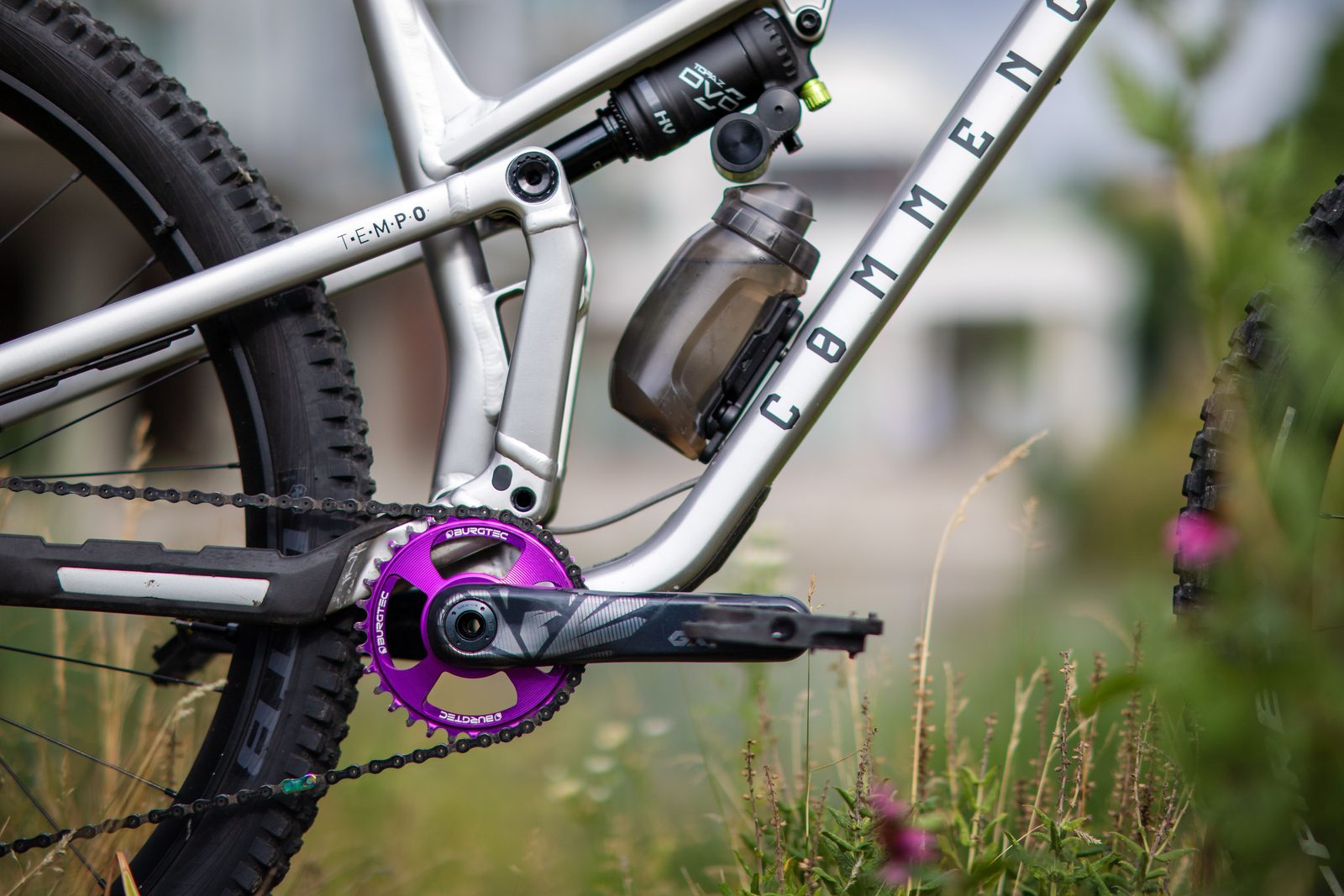 Close-up of a silver full-suspension mountain bike frame, crankset and water bottle in a meadow.